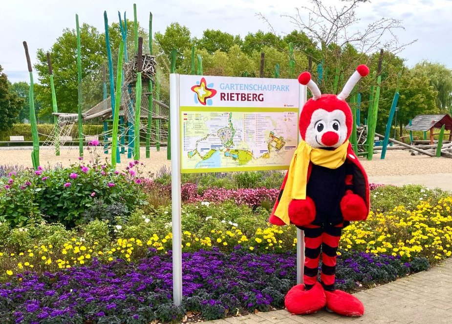 Maskottchen Rieti steht im Gartenschaupark neben einem großen Parkplan.