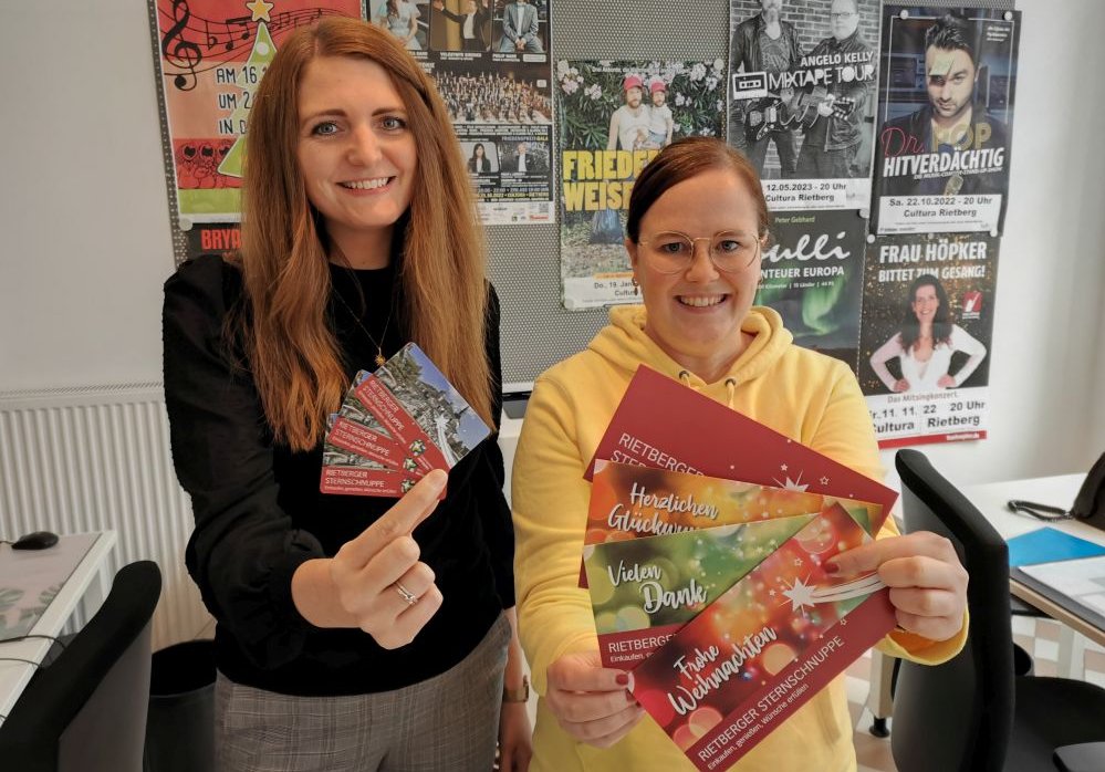 Touristik-Teamleiterin Lisa Jephcote und Mitarbeiterin Julia Wittreck stehen in der Touristikinformation und zeigen den Einkaufsgutschein Sternschnuppe samt zur Wahl stehenden Umschlägen mit den Aufdrucken "Herzlichen Glückwunsch", "Vielen Dank" und "Frohe Weihnachten".