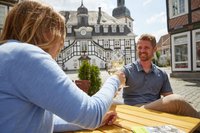 Zwei Menschen sitzen an einem Tisch und stoßen mit Weingläsern an. Im Hintergrund ist das Rietberger Rathaus zu sehen.