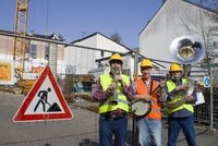 Drei Männer mit Instrumenten vor einer Baustelle, neben sich ein Baustellen-Schild. Sie tragen Warnwesten und auf dem Kopf Schutzhelme.