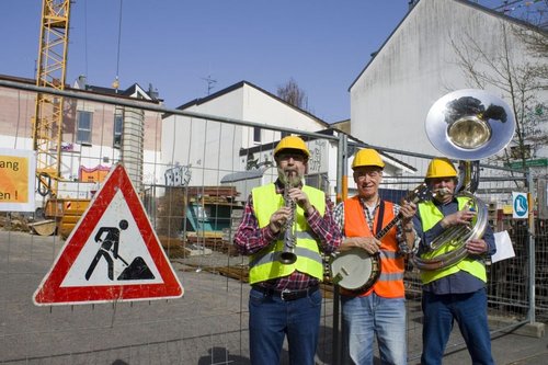 Drei Männer mit Instrumenten vor einer Baustelle, neben sich ein Baustellen-Schild. Sie tragen Warnwesten und auf dem Kopf Schutzhelme.