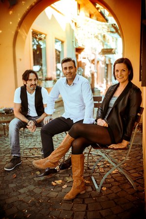 Salva, Desi und Daniele sitzen auf Stühlen vor einem großen Torbogen.