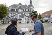 Zwei Radfahrer sind von hinten zu sehen. Sie halten einen Reiseführer in der Hand und schauen auf das Rietberger Rathaus (im Bildhintergrund).