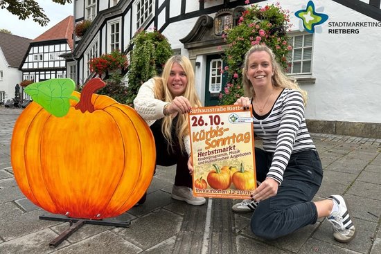 Bianca Piekatz (links) und Laura Hunke vom Stadtmarketing hocken mit Kürbissonntag-Plakat neben einem großen Kürbis aus Holz.