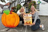 Bianca Piekatz (links) und Laura Hunke vom Stadtmarketing hocken mit Kürbissonntag-Plakat neben einem großen Kürbis aus Holz.