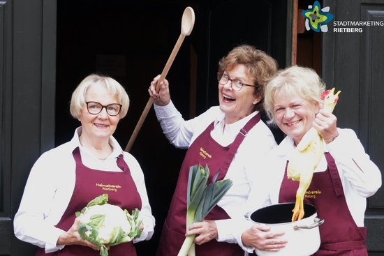 Die drei Gästeführerinnen, mit Schürzen bekleidet, in der Hand Blumenkohl, Kochtopf und Kochlöffel.